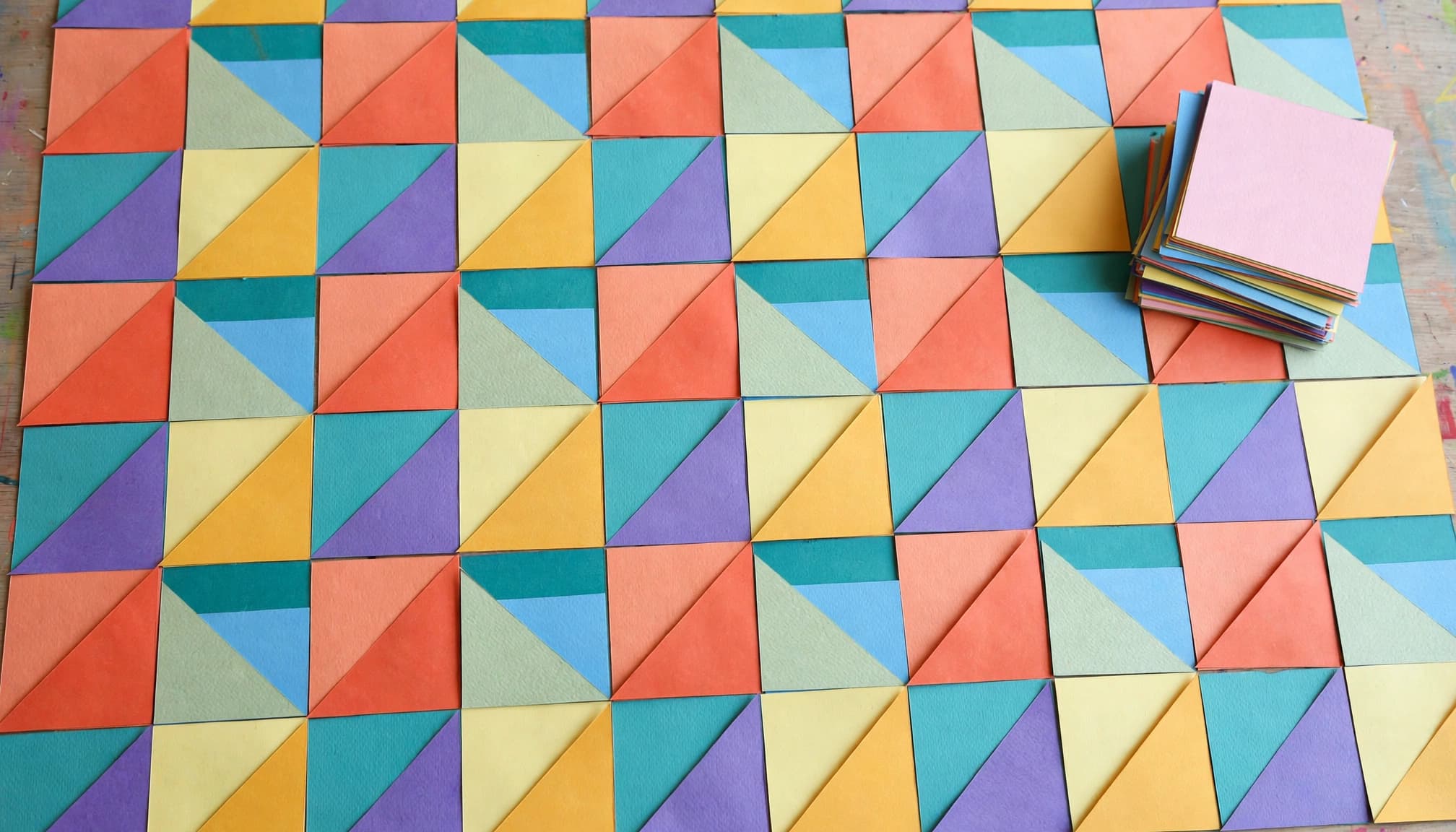 Colorful square and triangle paper tiles form a repeating pattern beside a small stack of shapes.